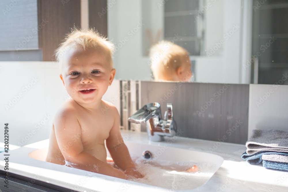 Wall mural Baby taking bath in sink. Child playing with foam and soap bubbles in sunny bathroom with window. Little boy bathing. Water fun for kids. Hygiene and skin care for children