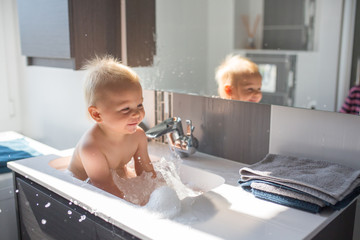 Baby taking bath in sink. Child playing with foam and soap bubbles in sunny bathroom with window. Little boy bathing. Water fun for kids. Hygiene and skin care for children © Tomsickova