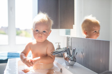 Baby taking bath in sink. Child playing with foam and soap bubbles in sunny bathroom with window. Little boy bathing. Water fun for kids. Hygiene and skin care for children - obrazy, fototapety, plakaty