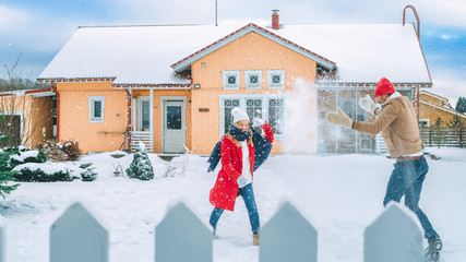 Young Beautiful Couple Throws Snowballs at Each other While Snow Falls. Happy Man and Woman Playing with Snow in the Yard of their Idyllic House. Family Enjoying Winter.