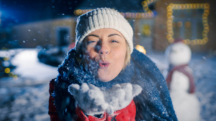 Happy Winter Evening, Laughing Young Woman Blows Snow From Her Hands. In the Background Idyllic House Decorated with Garlands for Christmas Eve. © Gorodenkoff