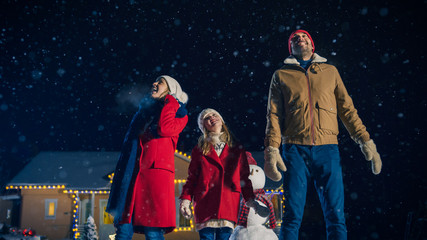 Happy New Year Celebration, Young Family of Three Standing in the Front Yard Looking into the Sky. In the Evening while Snow is Falling Father, Mother and Cute Little Daughter Look up in Wonder.