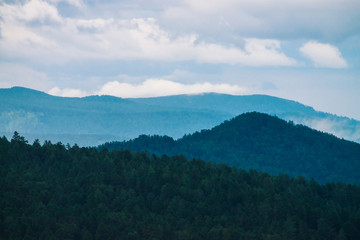 Obraz premium Gloomy green forest under blue mountains in fog on horizon in dusk. Atmospheric mystic evening mountain landscape of majestic nature.