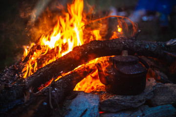 Boiling of tea in kettle on bonfire with large firewood. Tea drinking in open air. Active outdoor recreation. Camping in dusk. Romantic warm atmosphere in twilight on nature. Active rest.
