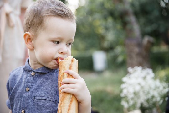 A picture of boy standing outside and holding baguette. He is biting a piece from it. Kid is looking backwards.