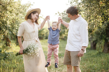 Fototapeta premium Happy family is walking together. Woman and man are holding their child on hands. They are looking at him. Woman is holding some flowers. They are happy. Child is smiling.