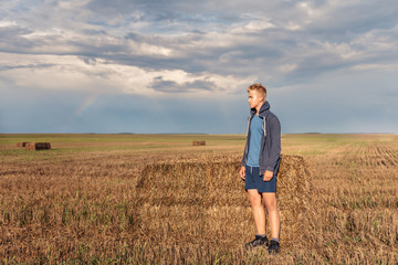 Naklejka na ściany i meble a wandering guy in a hoodie in a hood looks thoughtfully at a wheat field with a backdrop of the setting sun of a stormy sky