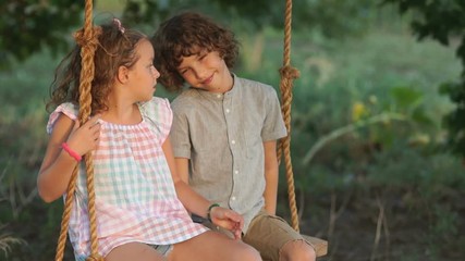 Loving boy and girl ride on a swing sitting next to each other. The boy kisses the girl, she hits him on the cheek, a slap in the face