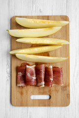 Melon slices with prosciutto on bamboo board over white wooden background, top view.