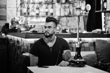 Stylish beard arabian man in glasses and black t-shirt smoking hookah indoor bar. Arab model having rest.