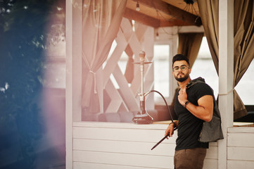 Stylish beard arabian man in glasses and black t-shirt smoking hookah outdoor. Arab model having rest.