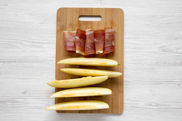 Fototapeta premium Melon slices with prosciutto on bamboo board over white wooden surface, top view. Overhead, flat lay, from above.