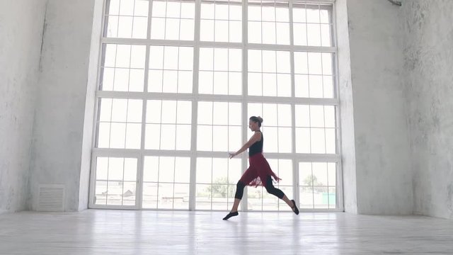 ballerina in a black bodysuit and a red skirt jumps high with a twine in the air in the Studio against a large light window. slow motion