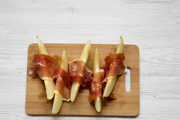 Melon slices wrapped in prosciutto on bamboo board over white wooden table, overhead view. From above, top view. Copy space.