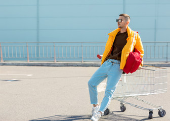 fashion guy in sunglasses and a yellow jacket jump in a cart from food in the supermarket parking