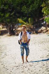 A stylish young man in jeans, a white shirt and sunglasses walking along the beach
