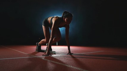 Slender young girl athlete is in position to start running in the pads on the track in slow motion