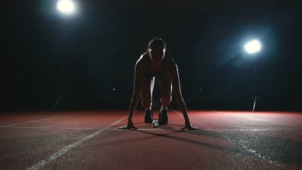 Slender young girl athlete is in position to start running in the pads on the track in slow motion