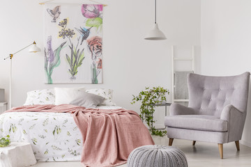 A sunny bedroom interior with a bed dressed in green pattern white linen and a peach blanket. Gray comfortable armchair beside the bed and a textile print of flowers and birds above. Real photo.