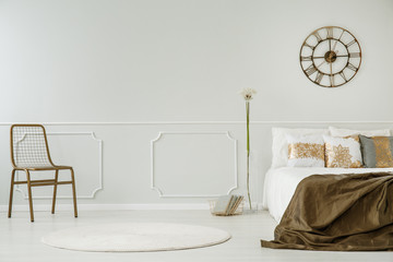 Gold clock above bed with blanket in white bedroom interior with chair and dandelion. Real photo
