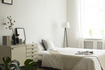 A white and beige color bedroom interior with a bed, a drawer cabinet and a lamp. Pillows, blanket and bedsheets on the bed. Real photo.