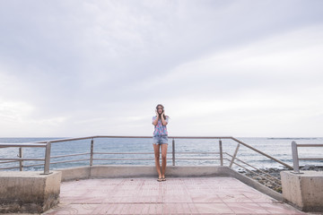 Full length portrait of beautiful young woman standing with the ocean and water blue in background and sky listening music with headphones. modern concept and leisure activity for nice girl