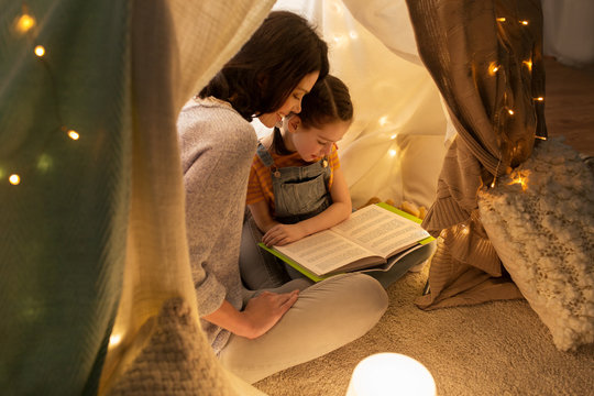 family, hygge and people concept - happy mother and little daughter reading book in kids tent at night at home