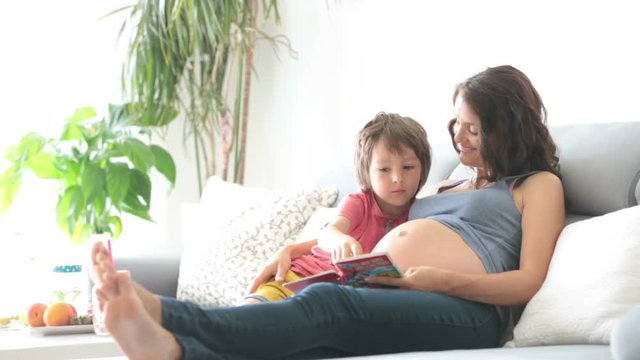 Young pregnant woman, reading a book at home to her boy, eating fruits, hugging and laughing