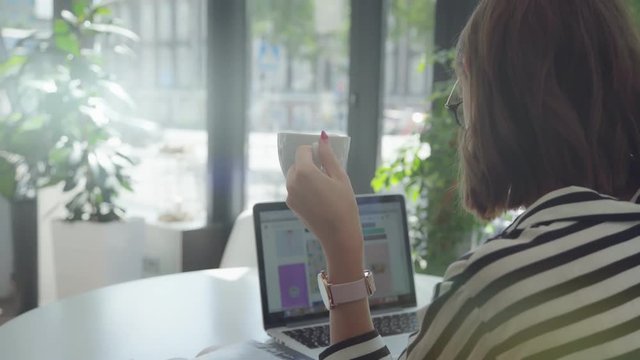 Taken over the left shoulder: the girl works behind the laptop and holds a cup of coffee in her hand. Only the back and screen of the laptop. The concept of working with a laptop, online, the Internet