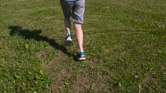 Boy runs fast on the green grass, close-up legs in sandals.
