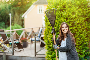 Smiling young woman with garden hose watering her home backyard with flowers, plants and...