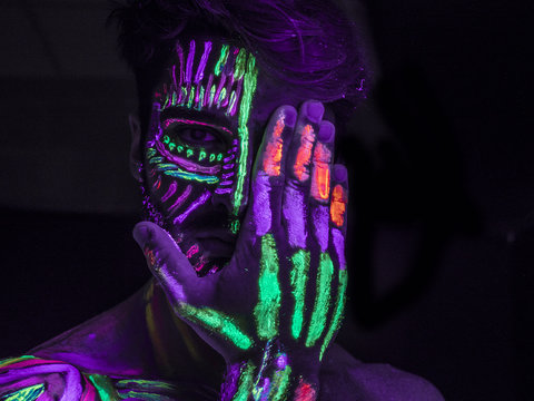 Headshot of young man painted in fluorescent paint on face and muscular torso, in studio shot with UV light