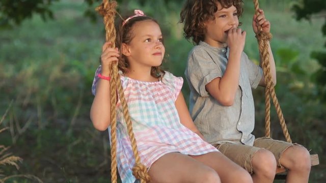 Couple of happy kids hugging and swinging on wooden swing at golden sunset. Brother embracing little sister on a swing. Young love between boy and girl on romantic summer evening