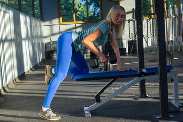 Blonde beatifull girl in the blue leggings and green t-shirt smile and push weight on the bench on the background of the composition of sports equipment and windows with trees in the open gym