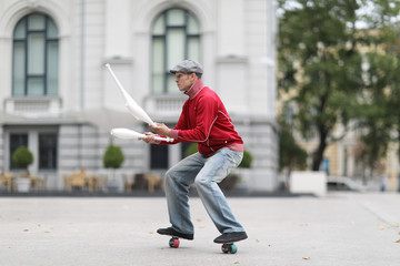 A man in a cap juggles with clubs, standing on small wheels on the street of a European city