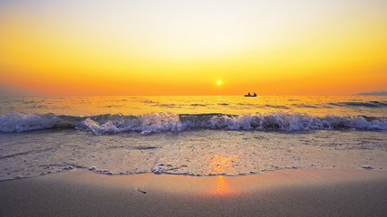 Small boat of refugees sail on sea horizon against sunset seen from beach splashed by waves