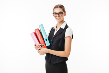 Young beautiful woman in black vest and eyeglasses happily looking in camera while holding colorful folders in hands over white background