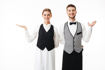 Young smiling waiter and pretty waitress in white shirts and vests happily looking in camera while...