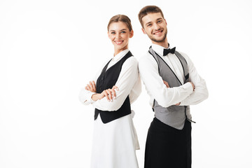 Young smiling waiter and waitress in white shirts and vests sstanding back to back while joyfully...