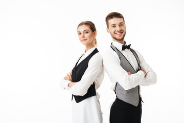 Young smiling waiter and beautiful waitress in white shirts and vests sstanding back to back while happily looking in camera with arms folded over white background - Powered by Adobe