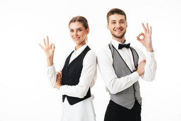 Young smiling waiter and waitress in white shirts and vests sstanding back to back happily looking...