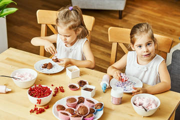 two beautiful sisters in white dresses decorate and eat delicious cupcakes at a wooden table