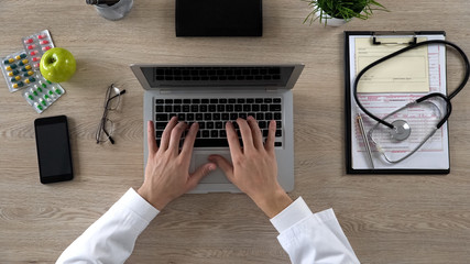 Medical worker typing on laptop, keeping electronic medical records, top view