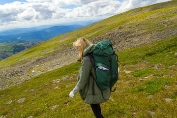 Young girl travel with big green backpack go down the hill on the rocks towards the green fields in...