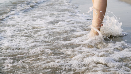 Closeup of female barefoot legs walking on sea coast being splashed by waves, cinematic steadicam shot