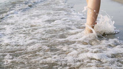 Fototapeta premium Closeup of female barefoot legs walking on sea coast being splashed by waves, cinematic steadicam shot