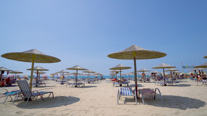 Summer beach resort bar lounge area with wooden easy chairs and straw parasols lined up next to the sea water