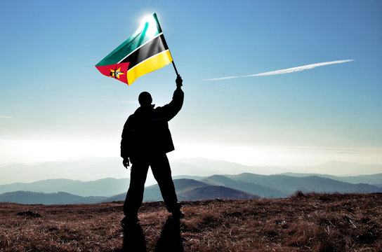 Successful silhouette man winner waving Mozambique flag on top of the mountain peak