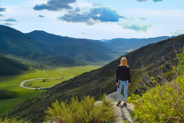 Fototapeta premium A young beautiful blonde girl stands on the edge of a cliff in the mountains of Altai on a bright sunny day, below is a green fresh grass and an asphalt road with a sharp turn leaving behind a rock