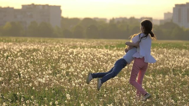 Two happy sisters hugged circling on the glade of dandelions against the background of city buildings outdoors. Freedom and carefree. Happy childhood.